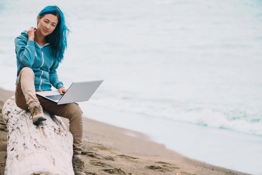 Freelancer young beautiful woman sitting on beach near the sea with laptop and looking at camera. Space for text in right part of the image