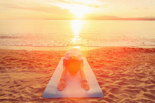 Young woman doing yoga exercise on sand beach near the sea at sunset in summer, woman stretching her back