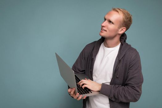 Side profile photo shot of handsome blonde man holding computer laptop typing on keyboard thinking about something wearing white t-shirt and grey sweater looking at netbook isolated over blue background. Free space