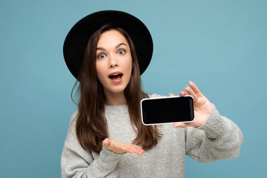 Portrait photo shot of beautiful amazing young woman wearing black hat and grey sweater holding phone showing smartphone isolated on background.Mock up, cutout