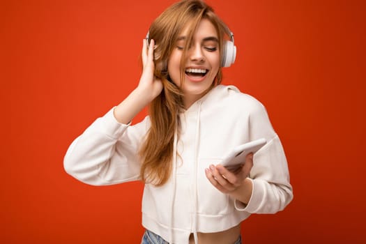 Photo of beautiful joyful smiling young woman wearing stylish casual clothes isolated over background wall holding and using mobile phone wearing white bluetooth headphones listening to music and having fun looking to the side.
