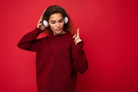 Photo of beautiful happy smiling young brunette curly woman wearing dark red sweater isolated over red background wall wearing white bluetooth headphones listening to music and having fun. copy space