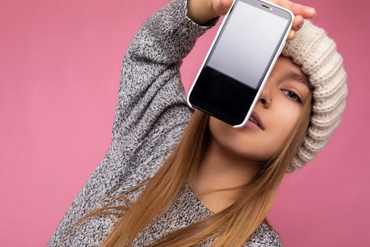 Photo of beautiful sexy young woman good looking wearing casual stylish outfit standing isolated on background with copy space holding smartphone showing phone in hand with empty screen display for mockup looking at camera.