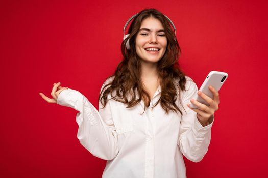 Attractive positive smiling young woman wearing stylish casual outfit isolated on colourful background wall holding and showing mobile phone with empty screen for cutout wearing white bluetooth headphones and having fun looking at camera.