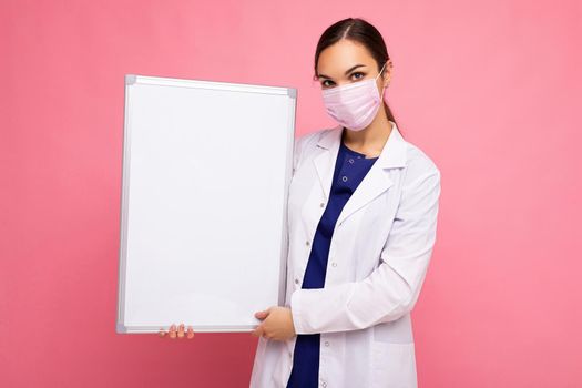 Woman doctor wearing a white medical coat and a mask holding blank board with copy space for text isolated on background. Coronavirus concept.