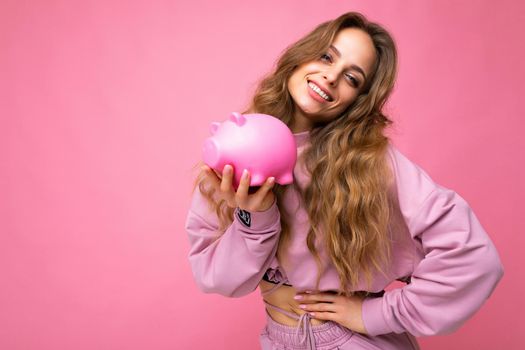 Portrait photo of happy positive smiling young beautiful attractive woman with wavy long blonde hair with sincere emotions wearing trendy pink hoodie isolated over pink background with copy space and holding pink pig penny bank. Money box concept.