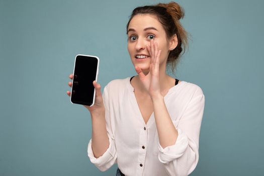 Beautiful positive young blonde curly woman wearing casual white shirt and isolated over blue background wall holding phone and showing smartphone with empty dcreen for mockup looking at camera and saying secret.