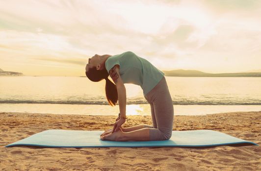 Young woman doing yoga exercise on sand beach on background of sea at sunset in summer