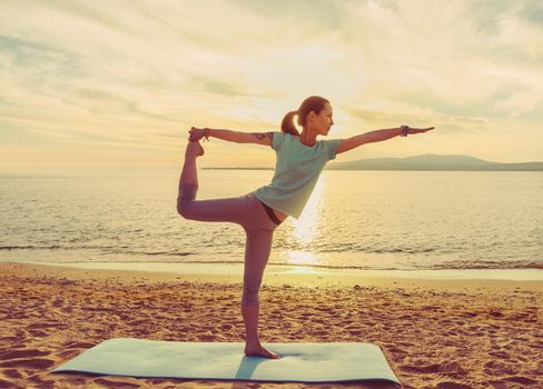 Young woman practicing yoga on mat on sand beach near the sea on sunset, woman in stretching balance pose
