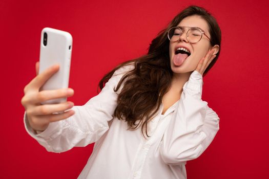 Closeup photo of beautiful positive young brunette woman wearing white shirt and optical glasses isolated over red background holding in hand and using mobile phone looking at gadjet screen and showing tongue.
