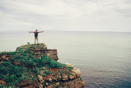 Happy traveler man standing with raised arms on peak of rock and enjoying view of sea