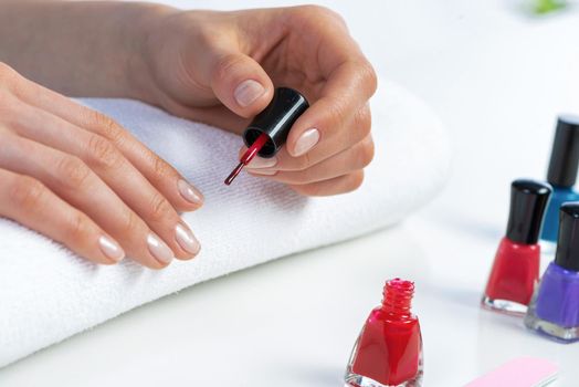 Woman giving herself elegant manicure at home. Closeup of beautiful female hand applying red nail polish. Colorful nail polish bottles on table. Nail care and beautician procedure. Stylish nail art