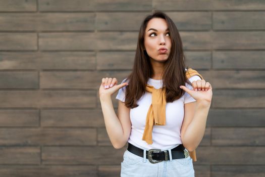 Photo of emotional positive happy joyful pretty young brunette woman in trendy outfit. Beautiful attractive female person standing outside in the street and showing sincere emotions and pointing at yourself.