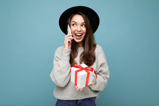 Beautiful happy young brunette woman isolated over colourful background wall wearing stylish casual clothes holding gift box and looking to the side.