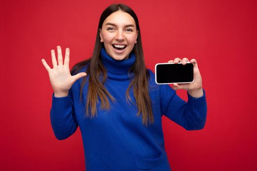 Closeup portrait photo shot of charming happy young brunette woman wearing casual blue sweater isolated over red background with empty space holding in hand mobile phone and showing smartphone with empty screen for mockup looking at camera and showing open palm and five fingers.