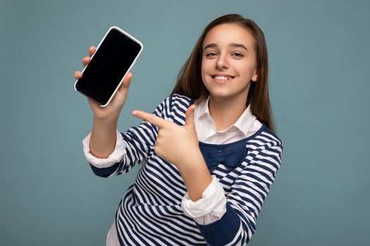 Photo of beautiful smiling girl good looking wearing casual stylish outfit standing isolated on background with copy space holding smartphone showing phone in hand with empty screen display for mockup pointing at gadjet looking at camera.