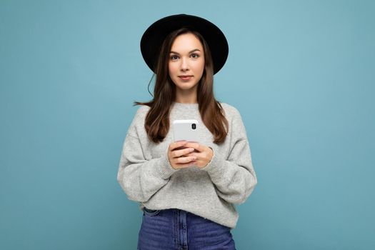 young brunette woman wearing black hat and grey sweater holding smartphone looking at camera isolated on background.