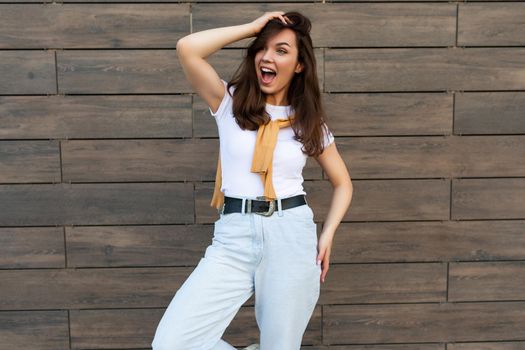 Portrait of successful smiling joyful happy young brunet woman wearing casual white t-shirt and jeans with yellow sweater poising near brown wall in the street and having fun.