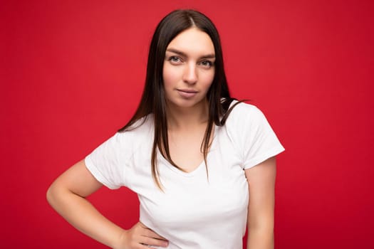Photo of self-confident adult beautiful brunette female person with sincere emotions wearing casual white t-shirt for mockup isolated over red background with copy space.