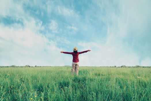 Young woman in straw hat standing with raised arms on meadow in summer. Concept of happiness and carefree