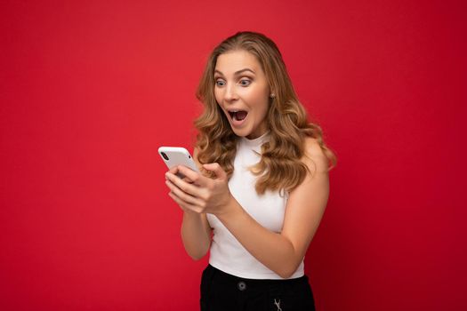 Photo shot of attractive shocked amazed positive good looking young woman wearing casual stylish outfit poising isolated on background with empty space holding in hand and using mobile phone messaging sms looking at smartphone display screen.