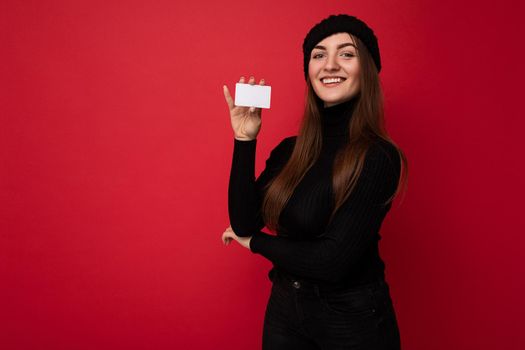 Photo of beautiful positive young brunette woman wearing black sweater and hat isolated on red background holding credit card looking at camera. copy space