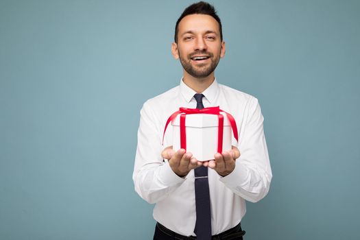 Photo shot of handsome positive brunette unshaven young man isolated over blue background wall wearing white shirt holding white gift box with red ribbon and looking at camera.