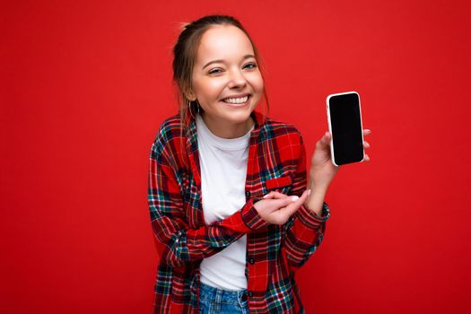 Photo of beautiful smiling young woman good looking wearing casual stylish outfit standing isolated on background with copy space holding smartphone showing phone in hand with empty screen display for mockup pointing at gadjet looking at camera.