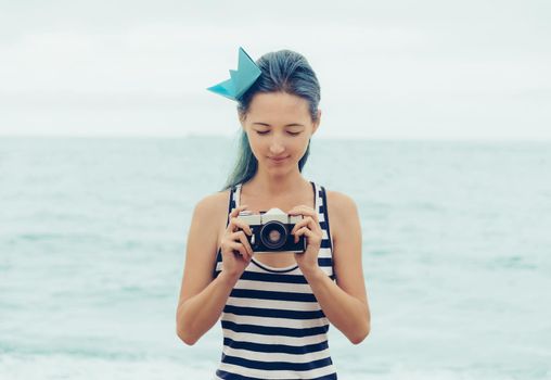 Attractive young woman in costume of sailor and origami ship in hairstyle standing with old photo camera on background of sea in summer