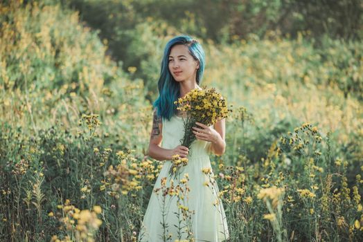 Beautiful young woman with blue hair and bouquet walking on flower field in summer