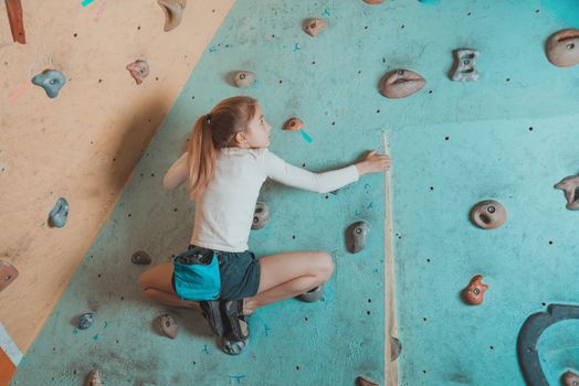 Climber little girl exercises in gym. Climber girl sitting on artificial boulders in pose of frog