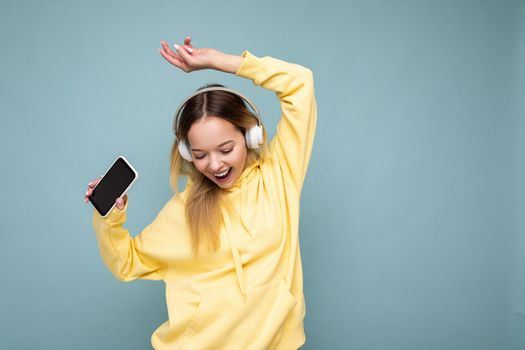 Photo of beautiful joyful smiling young woman wearing stylish casual clothes isolated over background wall holding and using mobile phone wearing white bluetooth headphones listening to music and having fun looking down.