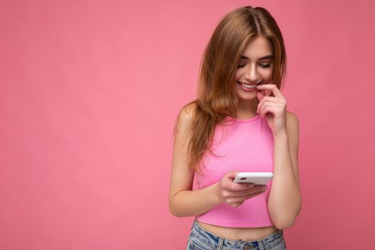 Photo shot of sexy attractive positive good looking young woman wearing casual stylish outfit poising isolated on background with empty space holding in hand and using mobile phone messaging sms looking at smartphone display screen.
