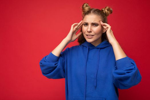 Photo of tired weary adult beautiful blonde woman with two horns with sincere emotions wearing hipster bright blue hoodie isolated over red background with copy space and having headache. Pain concept.