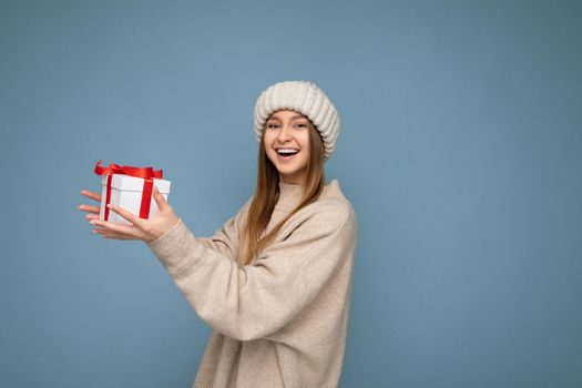 Shot of attractive positive smiling young dark blonde woman isolated over colourful background wall wearing everyday trendy outfit holding new year gift box with red ribbon and having fun looking at camera. Copy space
