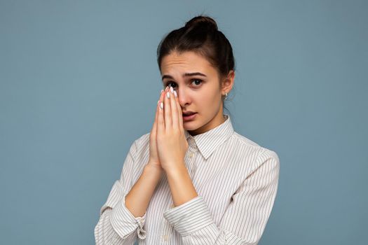 Photo of young attractive brunette woman with sincere emotions wearing white shirt isolated over blue background with empty space and holding hand together in pray.