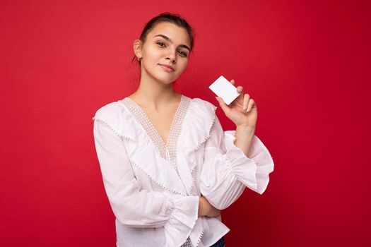 Beautiful happy self-confident young dark blonde woman wearing white blouse isolated over red background holding credit card looking at camera.