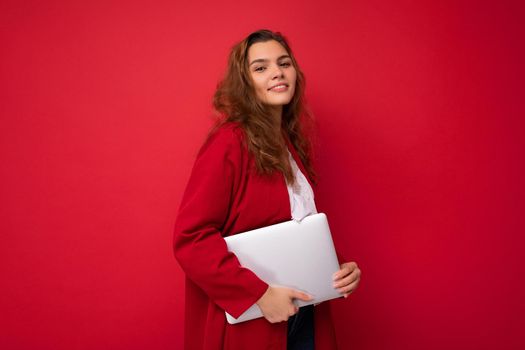 side profile photo of smiling beautiful charming pretty young lady holding laptop isolated over red wall background looking at camera.