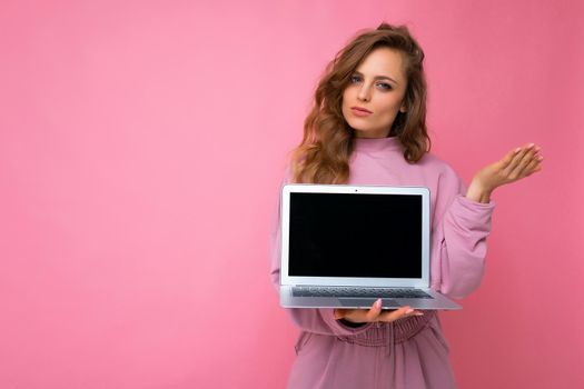 Close-up portrait of beautiful young dark blond curly hair woman holding computer laptop with empty copy space looking at camera and thinking about something having doubts posing hand isolated on pinl background.
