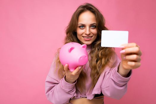 Happy positive smiling young beautiful charming woman with wavy long light hair with sincere emotions wearing stylish pink hoodie isolated over pink background with copy space, holds pink piggy box and credit card. Bank concept.