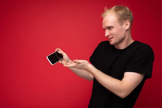 handsome positive happy blonde young male person wearing black t-shirt standing isolated on red background with copy space holding smartphone showing phone in hand with empty display for mockup pointing at gadjet looking at gadjet.