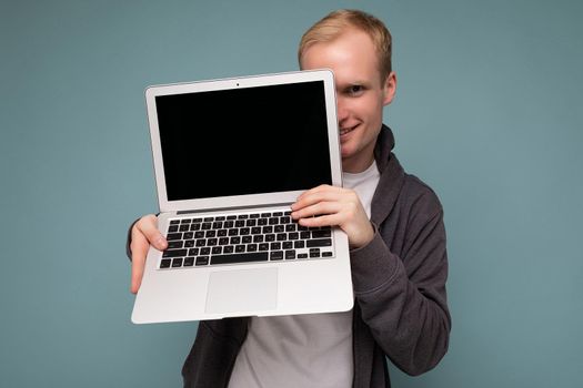 Photo shot of handsome smiling blonde man holding computer laptop with empty monitor screen with mock up and copy space wearing white t-shirt and grey sweater looking at camera isolated over blue background.