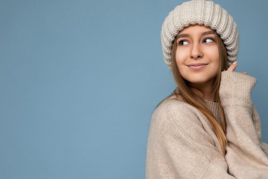 Shot of beautiful sexy smiling adult blonde woman standing isolated on blue background wall wearing beige warm sweater and winter beige hat looking to the side and enjoying. Empty space, copy space