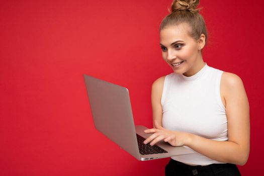 Side profile photo shot of beautiful overjoyed blond young woman with gathered hair wearing white t-shirt using computer laptop typing on keyboard looking at netbook monitor screen isolated over red wall background. Copy space