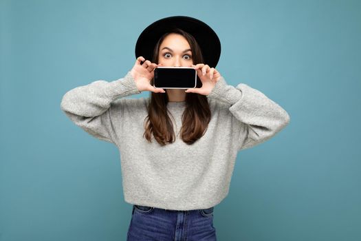 Photo of Beautiful positive young female person wearing black hat and grey sweater holding mobilephone showing smartphone isolated on background looking at camera.Mock up, cutout, empty space