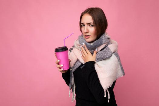 Photo shot of beautiful young brunette woman wearing black sweater and warm scarf isolated over pink background holding paper coffee cup for mockup drinking hot beverage and having cold.