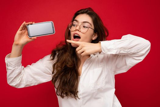 attractive shocked young brunet woman wearing white shirt and optical glasses isolated over red background holding in hand and showing mobile phone with empty screen for mockup looking at gadjet screen and pointing finger at gadjet display.