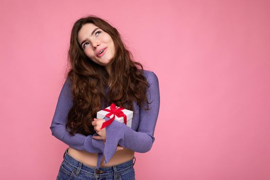 Pretty positive thoughtful brunet curly young female person isolated over pink background wall wearing purple blouse holding white gift box with red ribbon and looking up. Copy space