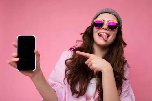 Positive attractive young brunette woman wearing stylish pink shirt grey hat and colourful sunglasses isolated over pink background holding in hand and showing mobile phone with empty display for mockup looking at camera pointing finger at gadjet and showing tongue.
