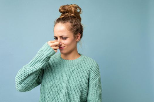 Portrait of young dissatisfied beautiful blonde woman with sincere emotions wearing casual blue pullover isolated on blue background with copy space and covers nose with hand, smells something awful and unpleasant.